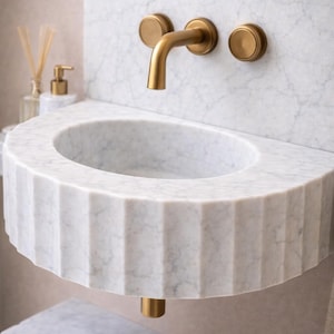 May include: A white marble sink with a fluted edge and gold-coloured tap and fixtures. The sink is oval-shaped and wall-mounted. The tap has a curved spout and two round handles. The design is modern and luxurious.