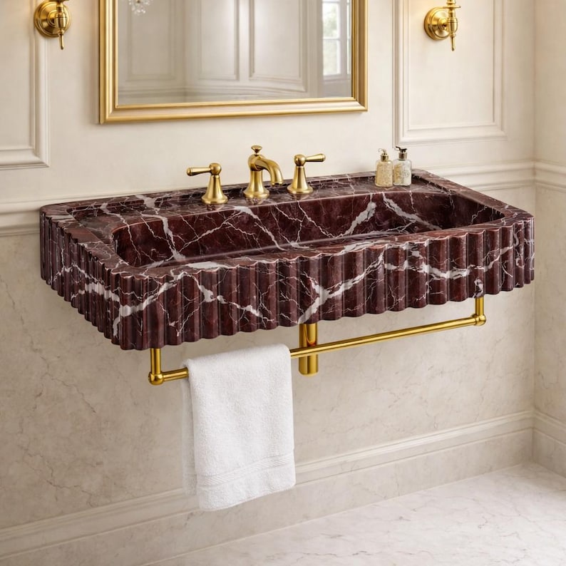 May include: A burgundy marble sink with white veining, gold fixtures, and a fluted edge. A gold towel bar holds a white towel. The sink has a rectangular shape and is mounted on a wall.