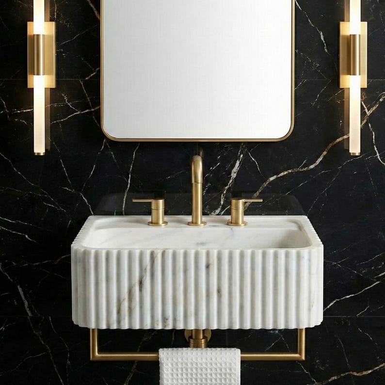 May include: A contemporary bathroom sink featuring a white marble basin and gold-coloured fixtures. The sink has a ribbed design and is mounted on a gold towel rail. A rectangular mirror with a gold frame hangs above the sink, set against a black marble wall.