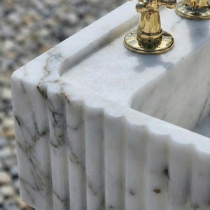 May include: A close-up of a white marble sink with gold-coloured taps. The sink has a rectangular shape with a fluted design on the front. The marble has grey and brown veining.