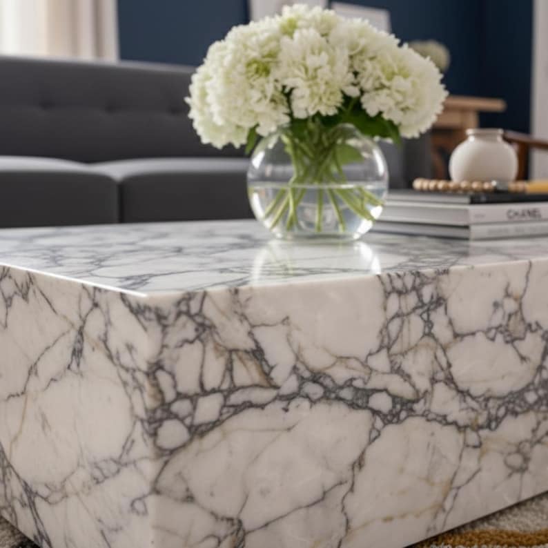May include: A white marble coffee table with gray veining. A clear glass vase holds white flowers. Books and a small white vase are on the table. The table is in a living room setting.