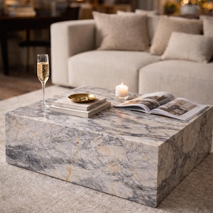 May include: A square marble coffee table with grey, white, and gold veining. A glass of champagne, a small gold dish, books, a lit candle, and an open magazine rest on the table. The table is on a light grey carpet.