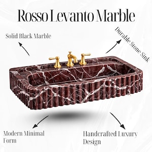 May include: A rectangular Rosso Levanto marble sink with fluted sides and gold-coloured taps. The durable stone sink has a modern, minimal form and handcrafted luxury design. The text "Rosso Levanto Marble" is at the top. The sink is made of solid black marble.