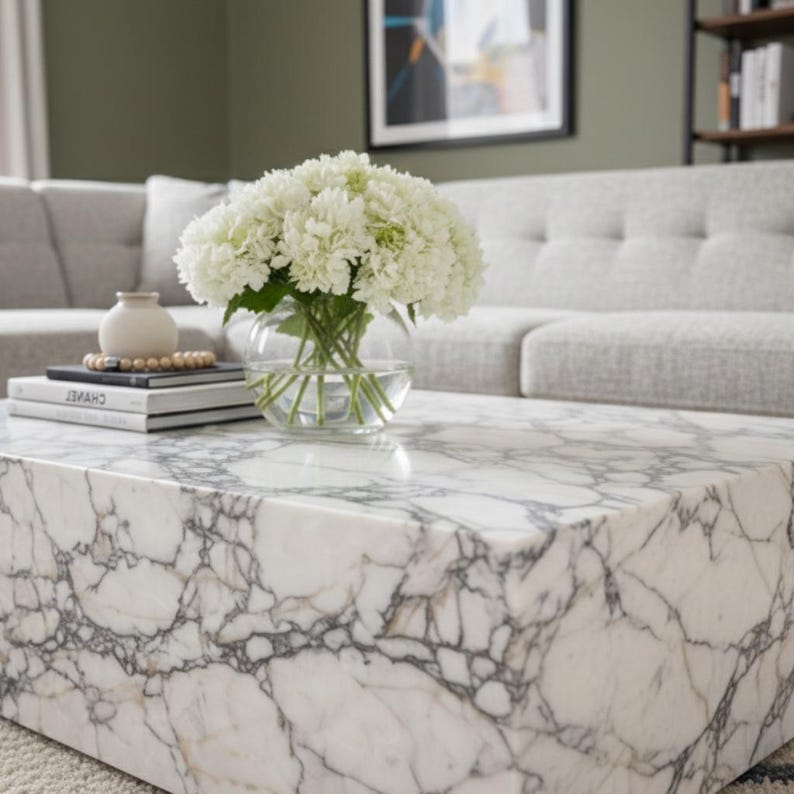 May include: A white marble coffee table with gray veining. A clear glass bowl with white flowers sits on top, along with a stack of books and a small vase. The table is in a living room setting with a gray sofa.
