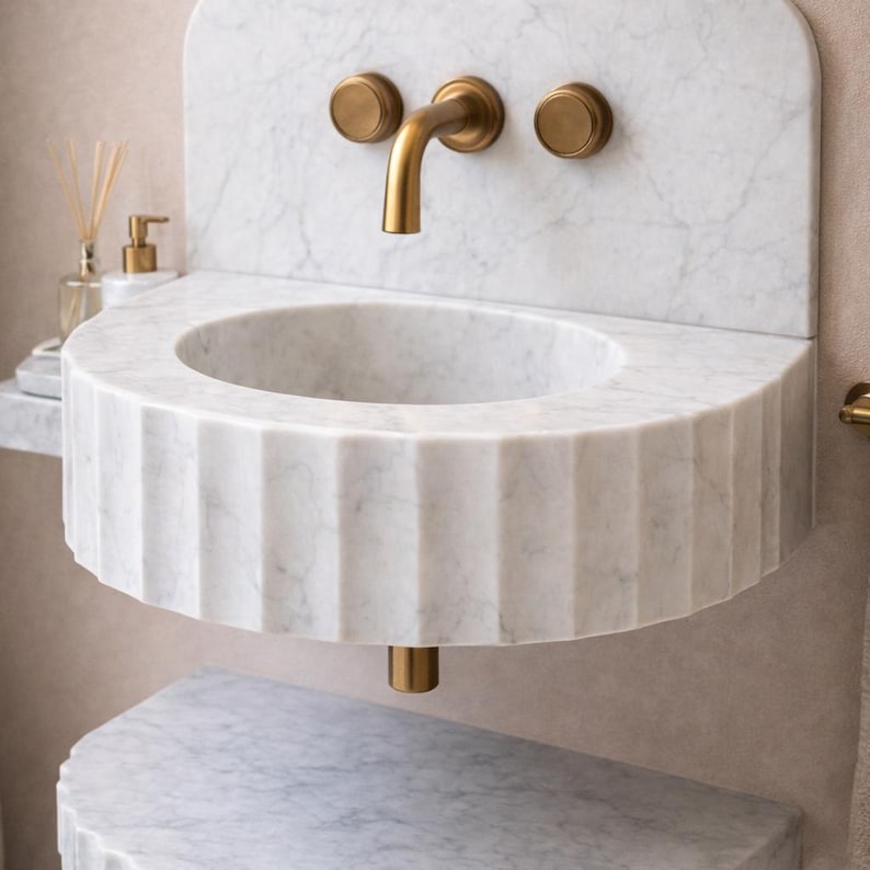 May include: A white marble sink with a fluted design, featuring a round basin and a brass tap. The sink is mounted on a marble wall with brass fixtures. A small shelf and a lower marble surface are also visible.