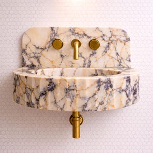May include: A marble wall-mounted sink with gold taps and fittings. The sink features a rounded basin and a rectangular backsplash. The marble is cream with dark grey and gold veining. The fittings have a brushed gold finish.