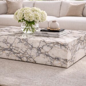 May include: A square marble coffee table with a white and gray veined pattern. A clear glass vase with white flowers sits on the table, along with books and decorative beads. The table is in front of a white sofa.