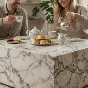 May include: A marble coffee table with a white and gray veined pattern. A tea set with floral designs, scones, and cookies are arranged on the table. A person is pouring tea from a silver teapot into a teacup.