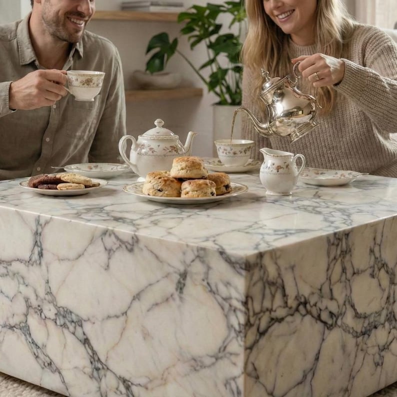 May include: A marble coffee table with a white and gray veined pattern. A tea set with floral designs, scones, and cookies are arranged on the table. A person is pouring tea from a silver teapot into a teacup.