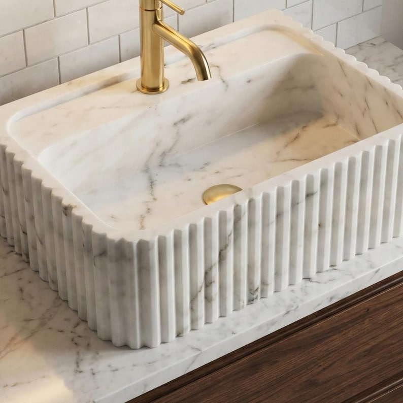 May include: A white marble sink with a fluted design and gold-coloured tap. The sink is rectangular and sits on a marble countertop, above a dark wood cabinet. The sink's dimensions are in cm.