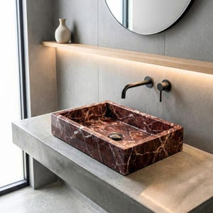 Op de afbeelding: Een rechthoekige wastafel van rood marmer met witte adering. De wastafel is verzonken in een betonnen aanrechtblad. Een zwarte kraan en armaturen zijn op de muur boven de wastafel gemonteerd. Een ronde spiegel en een kleine vaas zijn zichtbaar op de achtergrond.