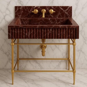 May include: A burgundy marble sink with a fluted front and gold fixtures. The sink has a rectangular basin and is mounted on a gold metal stand. The faucet and knobs are also gold. The background is a marble wall.