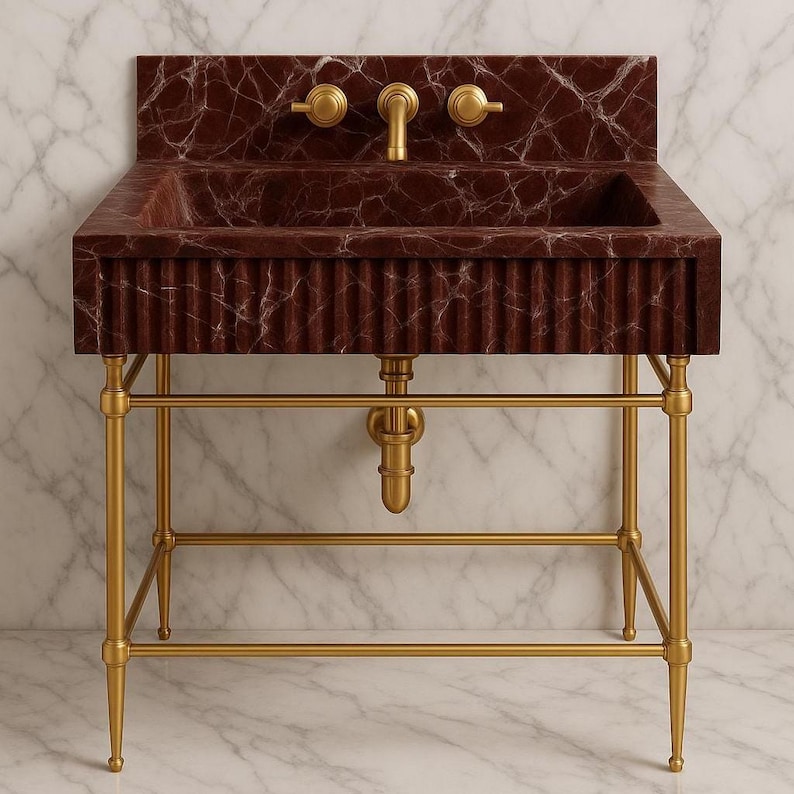 May include: A burgundy marble sink with a fluted front and gold fixtures. The sink has a rectangular basin and is mounted on a gold metal stand. The faucet and knobs are also gold. The background is a marble wall.
