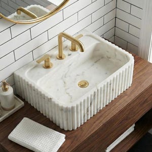 May include: A rectangular white marble sink with a fluted design, gold tap and matching fixtures. The sink is set on a dark wood vanity. A white waffle-weave towel and soap dispenser are visible.
