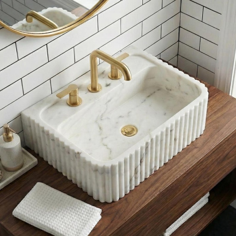 May include: A rectangular white marble sink with a fluted design, gold tap and matching fixtures. The sink is set on a dark wood vanity. A white waffle-weave towel and soap dispenser are visible.