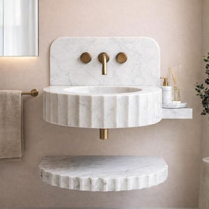 May include: A contemporary white marble washbasin with a fluted design and a matching shelf. The basin has gold-coloured taps and fittings. A towel rail and mirror are visible in the background.