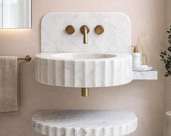 May include: A modern, white marble bathroom sink with a fluted basin and a matching shelf below. The sink features a gold-toned faucet and fixtures. A towel rack and mirror are visible in the background.