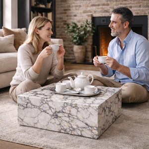 May include: A couple enjoys tea in a living room setting. A white marble coffee table holds a silver tray with a white teapot, sugar bowl, and teacups. The table is rectangular and features a gray and gold veined pattern. The couple is seated on the floor.