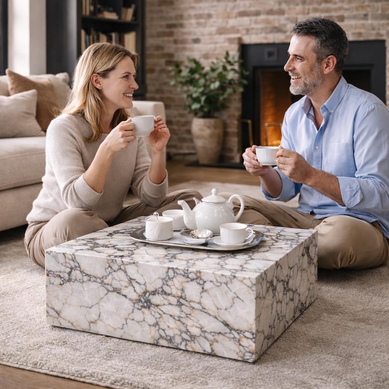 May include: A couple enjoys tea in a living room setting. A white marble coffee table holds a silver tray with a white teapot, sugar bowl, and teacups. The table is rectangular and features a gray and gold veined pattern. The couple is seated on the floor.