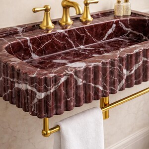 May include: A red marble bathroom sink with white veining and a fluted edge. The sink has gold-coloured taps and a matching towel rail. A white towel hangs from the rail. The sink is a luxurious bathroom fixture.