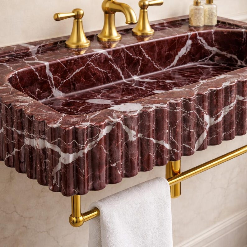 May include: A red marble bathroom sink with white veining and a fluted edge. The sink has gold-coloured taps and a matching towel rail. A white towel hangs from the rail. The sink is a luxurious bathroom fixture.