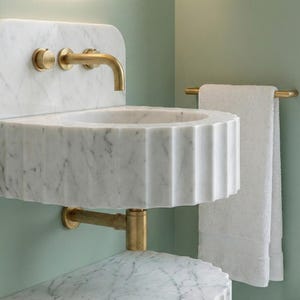 May include: A white marble sink with a fluted design and brass fixtures. The sink has a rounded shape and is mounted on a wall. A brass towel rail holds a white towel. The background is a light green colour.