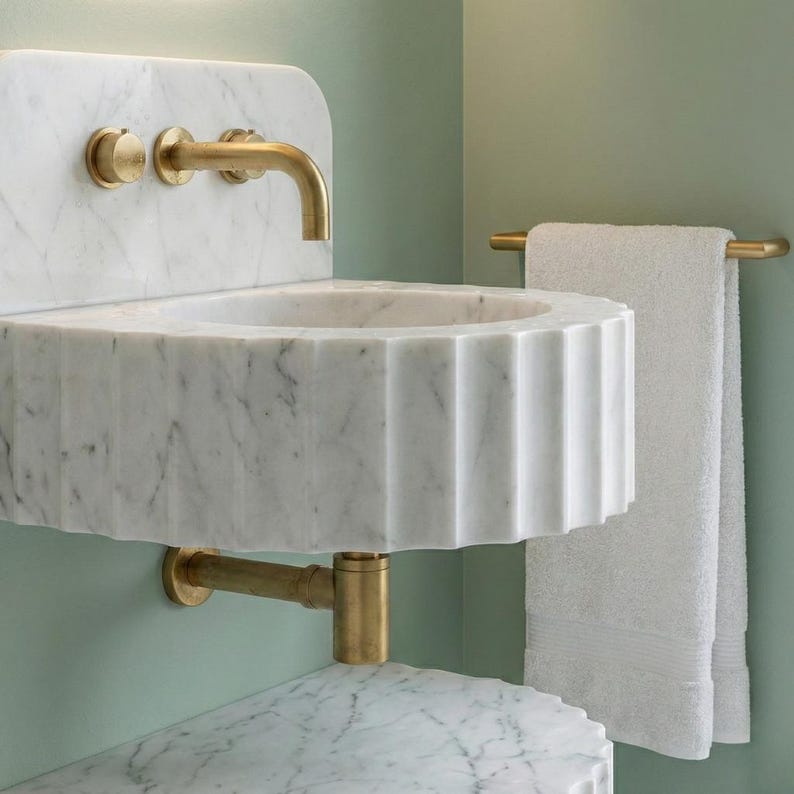 May include: A white marble sink with a fluted design and brass fixtures. The sink has a rounded shape and is mounted on a wall. A brass towel rail holds a white towel. The background is a light green colour.