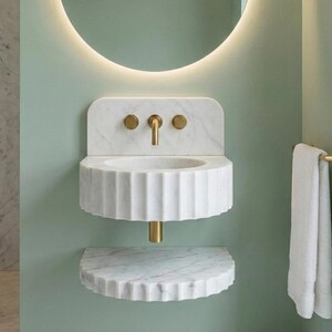 May include: A white marble wall-mounted sink with a fluted basin and a matching shelf. The sink has a gold tap and fixtures. The background is a light green wall with a circular mirror and a towel rail.