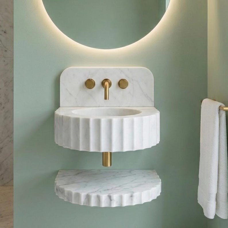 May include: A white marble wall-mounted sink with a fluted basin and a matching shelf. The sink has a gold tap and fixtures. The background is a light green wall with a circular mirror and a towel rail.