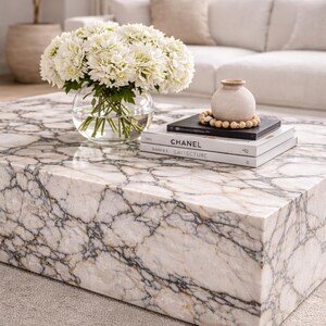 May include: A rectangular marble coffee table with a white and gray veined pattern. A clear glass vase holds white flowers. Books with the title "CHANEL" and "CRITECTURE" are stacked on the table, along with a small vase and wooden beads.