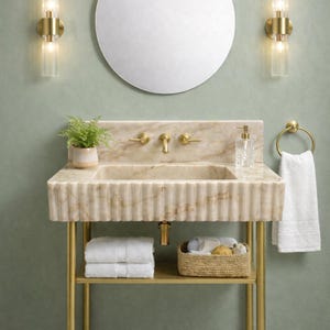 Puede incluir: Un lavabo de mármol con frente estriado, grifo de latón y cubeta integrada. La unidad cuenta con un estante inferior con dos toallas blancas dobladas y una cesta tejida, flanqueado por un toallero de aro y dos luces de pared.