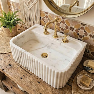May include: A white marble sink with a fluted design and gold taps. The sink is set on a wooden vanity. The taps and waste are gold. The background includes a round mirror and decorative tiles. A potted fern and soap dishes are also visible.