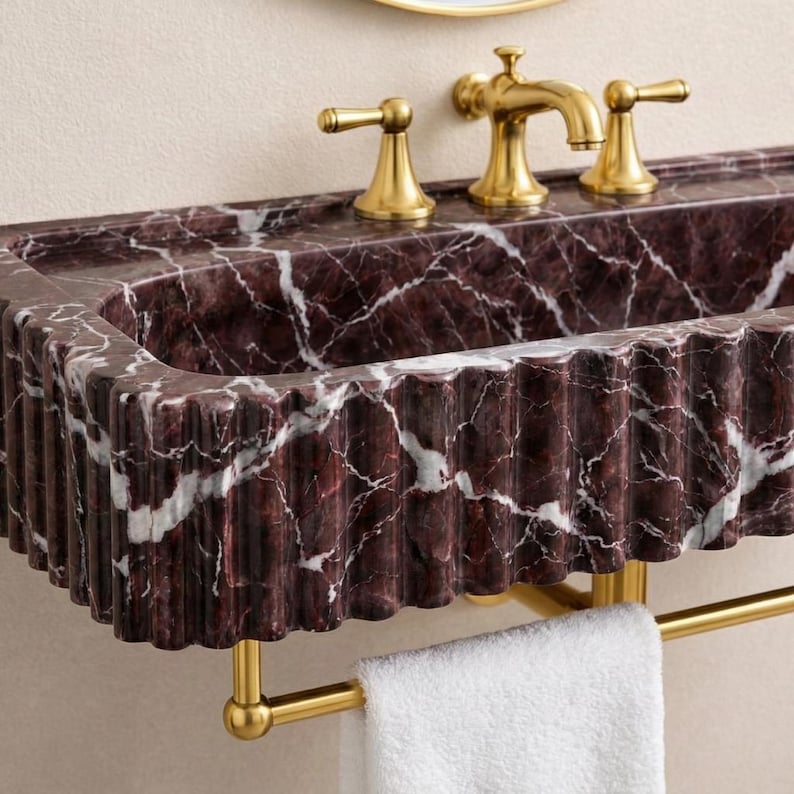 May include: A close-up of a bathroom sink with a dark red marble basin and gold fixtures. The basin has a fluted edge and is mounted on a wall. A gold faucet and two handles are visible, along with a gold towel rack and a white towel.