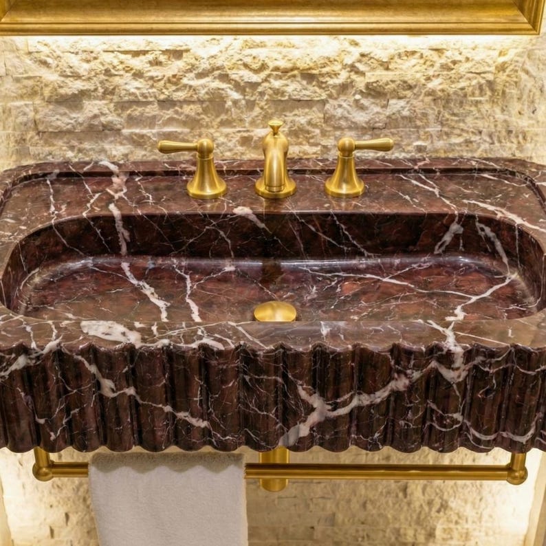 May include: A rectangular bathroom sink made of dark red marble with white veining. The sink has a scalloped edge and gold-coloured taps and drain. A gold towel rail is below the sink.