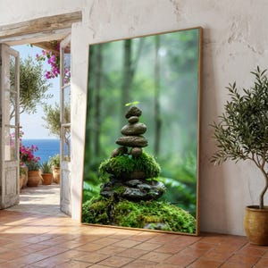 May include: A framed print featuring a balanced stack of stones covered in moss, with a small green sprout at the top. The background is a blurred forest scene. The print is displayed indoors near an open doorway.