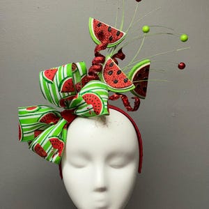 May include: A whimsical watermelon-themed headband. The headband features a large bow with green and white striped ribbon, adorned with watermelon slices. Red glitter accents and decorative sprigs complete the festive design.