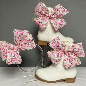 May include: White cowboy boots and a headband with large pink flamingo bows. The bows are made of a white fabric with a pink flamingo pattern. The boots have a low heel and a classic western design.