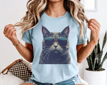 May include: Light blue t-shirt featuring a graphic of a cat wearing sunglasses. The cat illustration is detailed with shades of gray and black, and the sunglasses have a gold frame and blue lenses. The shirt has short sleeves.