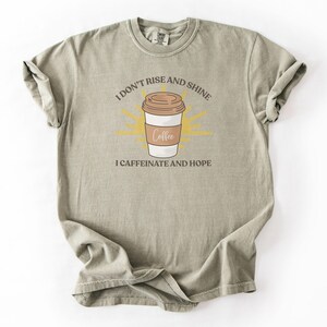 Peut inclure: T-shirt vert sauge avec le texte "I don't rise and shine, I caffeinate and hope." Le graphisme présente une tasse à café avec un couvercle marron et un motif en forme d'éclat de soleil jaune.