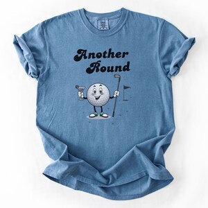 May include: Blue t-shirt with the text "Another Round" in black script. The shirt features a cartoon golf ball character holding a martini and a golf club. A golf flag is also in the design.