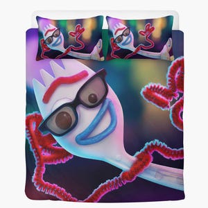 May include: Bedding set with a cartoon character design. The duvet cover and two pillowcases feature a white character with black glasses, a red pipe cleaner arm, and a blue smile. The background is a blurred mix of colours.