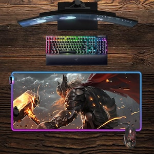 May include: A gaming setup featuring a curved monitor, a backlit keyboard with rainbow lighting, a mouse, and a large mousepad with a Thor graphic. The mousepad has a glowing border.