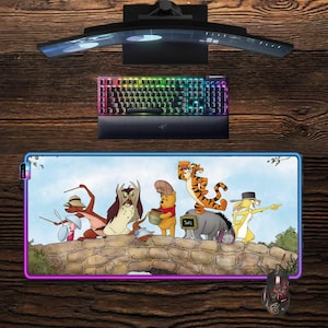 Pode incluir: Uma configuração de computador com um monitor curvo, um teclado retroiluminado e um rato. Um grande tapete de rato retangular exibe uma ilustração colorida de Winnie the Pooh e seus amigos em uma ponte. O tapete tem uma borda brilhante.