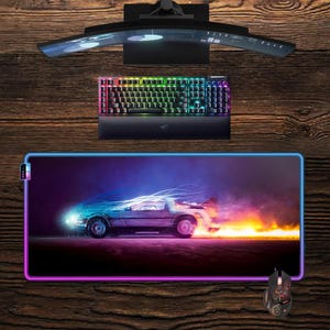 May include: A gaming setup featuring a curved monitor, a backlit keyboard, and a mouse. A large rectangular mousepad displays a futuristic car with neon lights and flames. The keyboard and mouse have colorful lights.