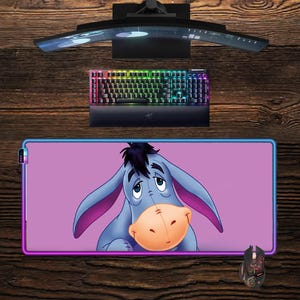 Pode incluir: Uma configuração de computador com um monitor curvo, um teclado retroiluminado e um rato. A peça central é um tapete de rato retangular com uma imagem de desenho animado de Eeyore de Winnie the Pooh. O tapete tem um fundo roxo e uma borda colorida.