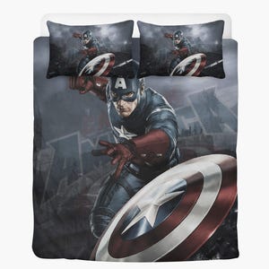 May include: Bedding set featuring Captain America in action, with his shield. The set includes a duvet cover and two pillowcases. The design is predominantly blue, red, and white, with a cityscape background. The pillowcases show Captain America in a similar pose.