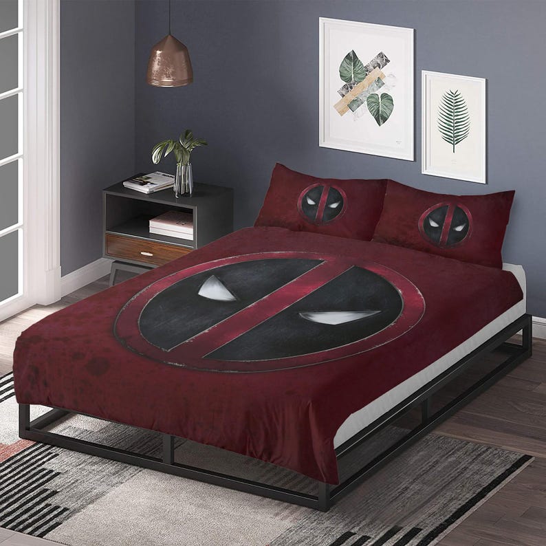 May include: A burgundy bed set featuring a large black and red Deadpool logo on the duvet cover and matching pillowcases. The bed is on a black metal frame, with a gray and white patterned rug at the foot.