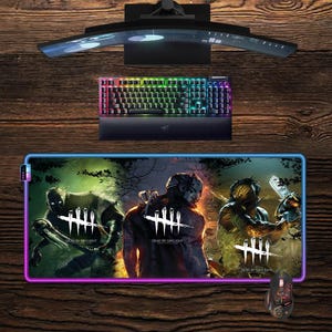 May include: A gaming setup featuring a curved monitor, a backlit keyboard, and a mouse. A large mousepad displays a graphic from "Dead by Daylight" with a blue and purple border. The keyboard has rainbow-colored keys.