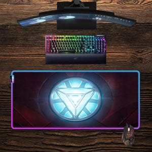 Puede incluir: Una configuración de juego con un monitor curvo, un teclado con retroiluminación arcoíris y una alfombrilla de ratón con un diseño de reactor de arco Iron Man brillante. También hay un ratón para juegos a juego. Los artículos están dispuestos sobre una superficie de madera.