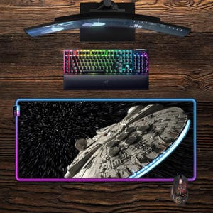 Könnte beinhalten: Gaming-Setup mit einem gebogenen Monitor, einer Tastatur mit Regenbogenbeleuchtung, einer Maus und einem großen Mauspad mit dem Millennium Falcon aus Star Wars. Auf einer Holzoberfläche.